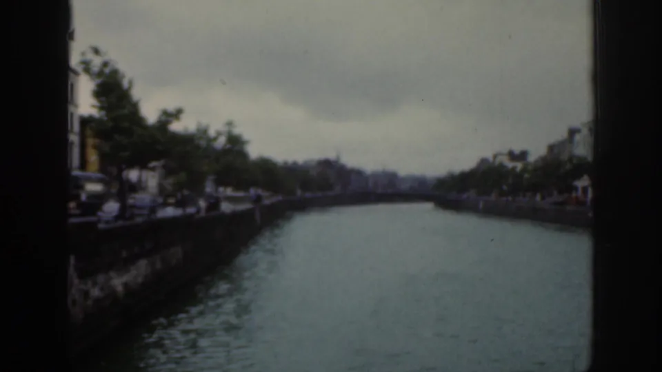 1961 Dublin home-movie view along the river, showing a stone quay, roadside vehicles, trees, and distant riverside buildings.