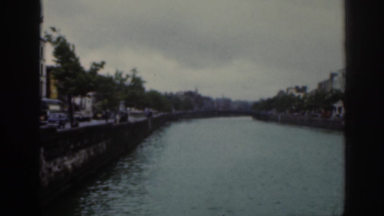 1961 Dublin frame looking down a river between stone embankments, with trees, buildings, muted color, and visible film borders.
