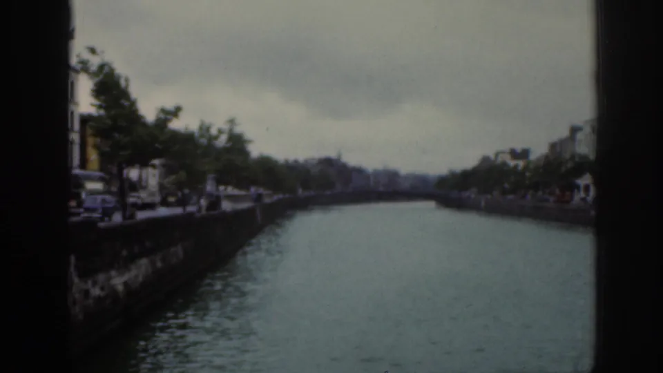 1961 Dublin frame looking down a river between stone embankments, with trees, buildings, muted color, and visible film borders.