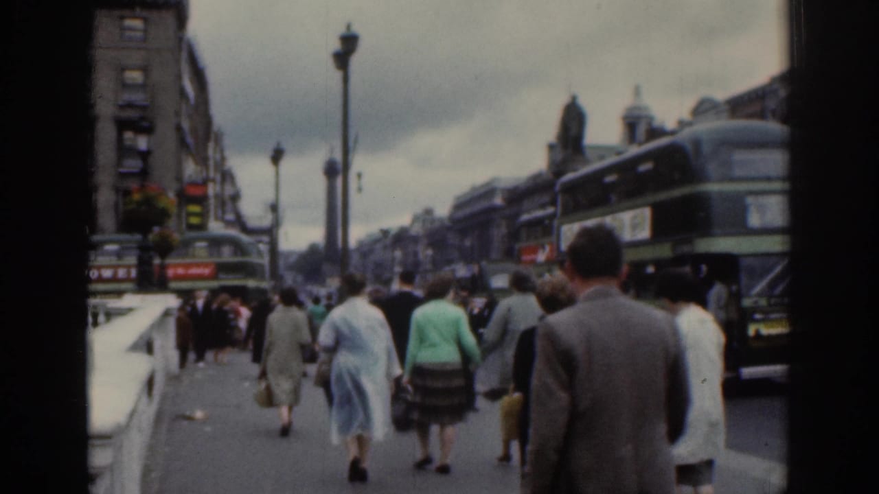 1961 Dublin street scene with pedestrians walking along a bridge or riverside pavement beside double-decker buses and older buildings.