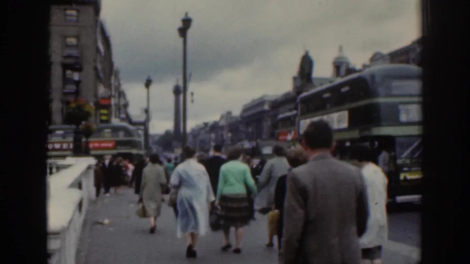 1961 Dublin street scene with pedestrians walking along a bridge or riverside pavement beside double-decker buses and older buildings.