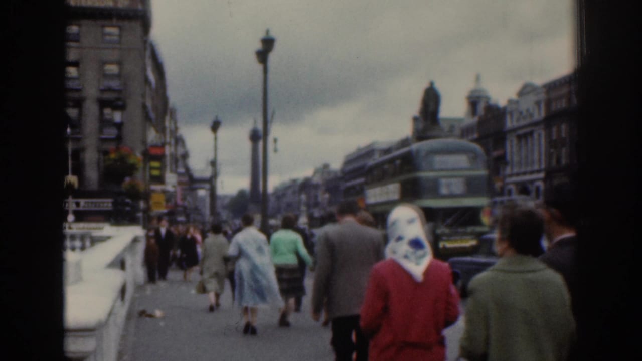 1961 Dublin home-movie frame showing pedestrians in period clothing walking toward buses, shopfronts, and street lamps.