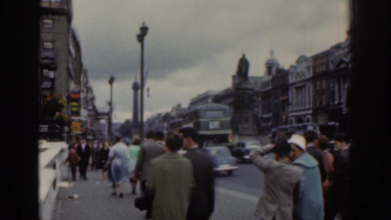 1961 Dublin street footage with pedestrians on the pavement, a vintage car in traffic, and a double-decker bus near older city buildings.