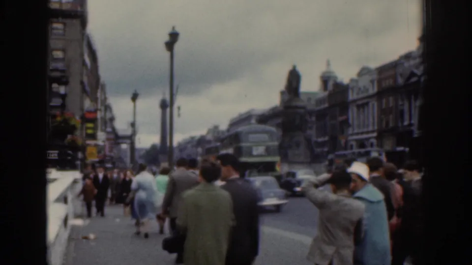 1961 Dublin street footage with pedestrians on the pavement, a vintage car in traffic, and a double-decker bus near older city buildings.