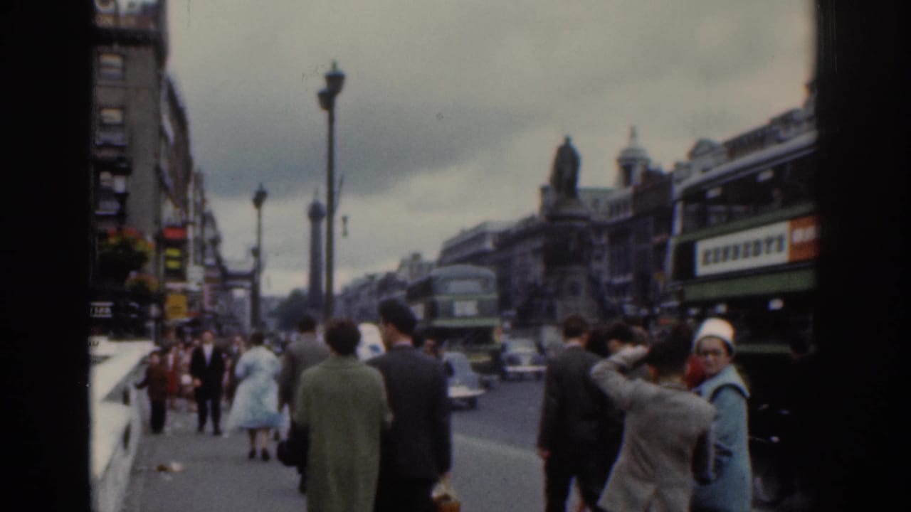 1961 Dublin street view with pedestrians in coats, green double-decker buses, classic cars, ornate lamp posts, and a distant statue.