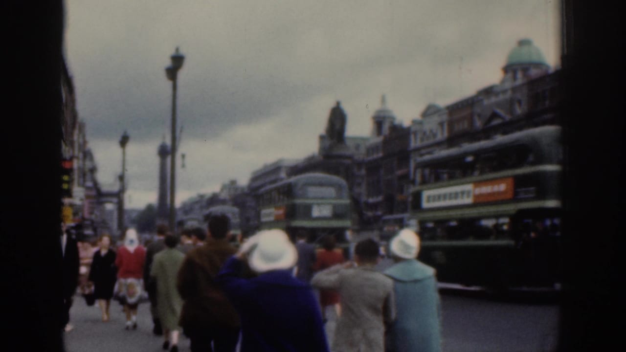 1961 Dublin street footage showing pedestrians from behind, light-colored hats, green buses, cars, lamp posts, and older city facades.