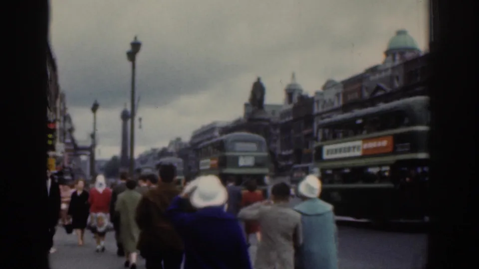 1961 Dublin street footage showing pedestrians from behind, light-colored hats, green buses, cars, lamp posts, and older city facades.