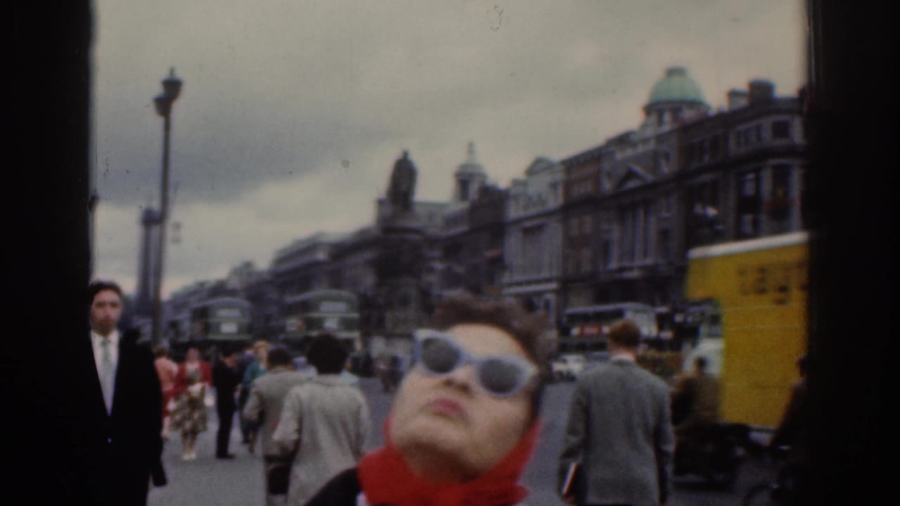 In a 1961 Dublin street view, a woman in sunglasses and a red headscarf looks up toward the camera as buses and pedestrians pass behind her.