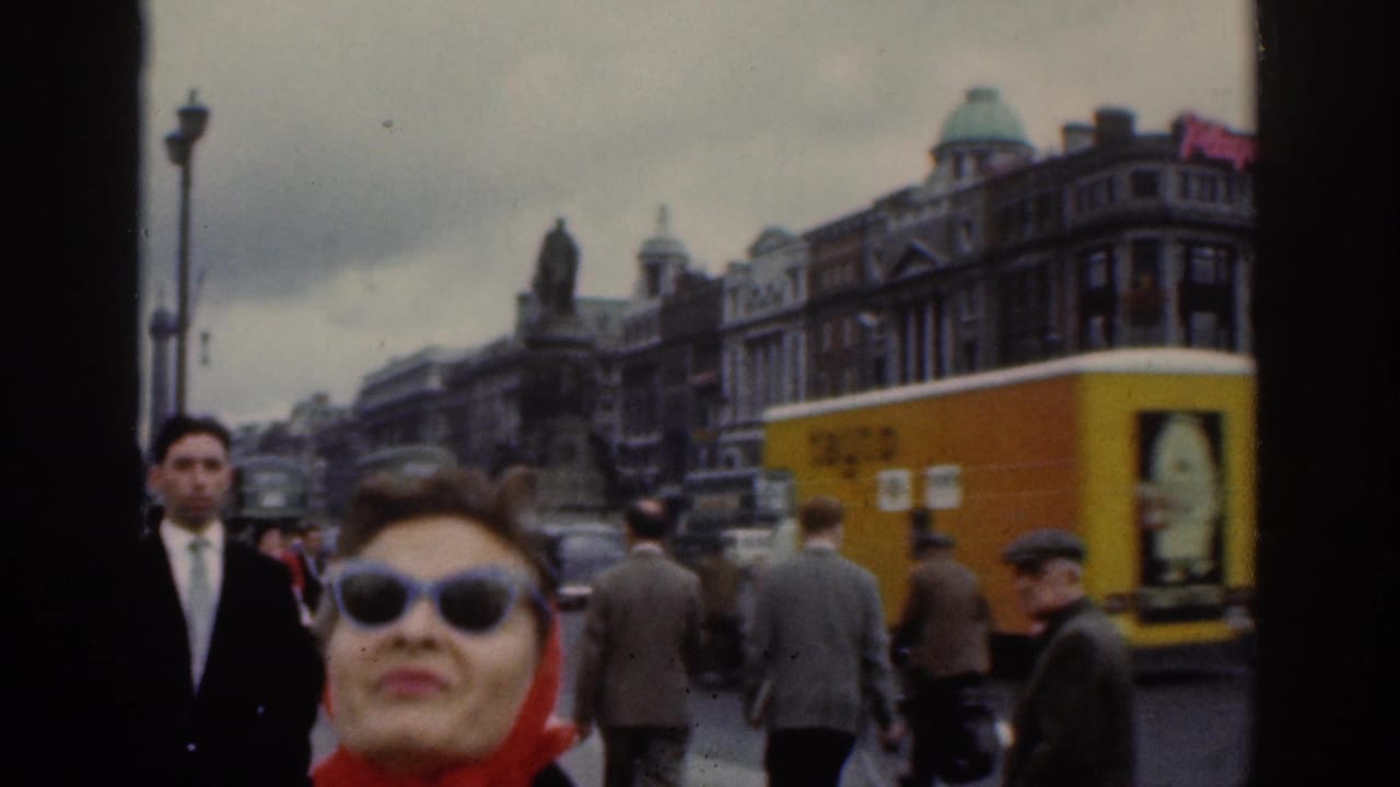 1961 Dublin street footage centers the woman in a red headscarf and sunglasses, with pedestrians, a yellow vehicle side, cars, and buildings behind her.