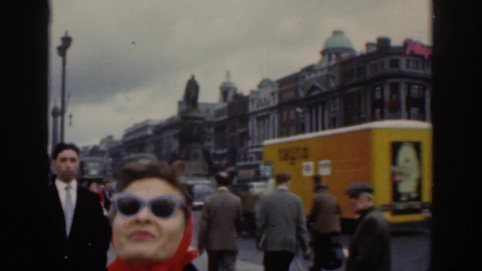 1961 Dublin street footage centers the woman in a red headscarf and sunglasses, with pedestrians, a yellow vehicle side, cars, and buildings behind her.