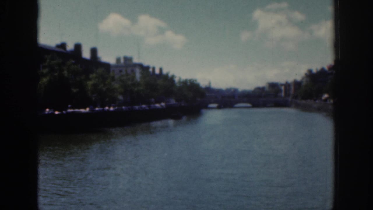 1961 Dublin river scene from a moving 8mm camera, with water leading toward a low bridge between shaded embankments.