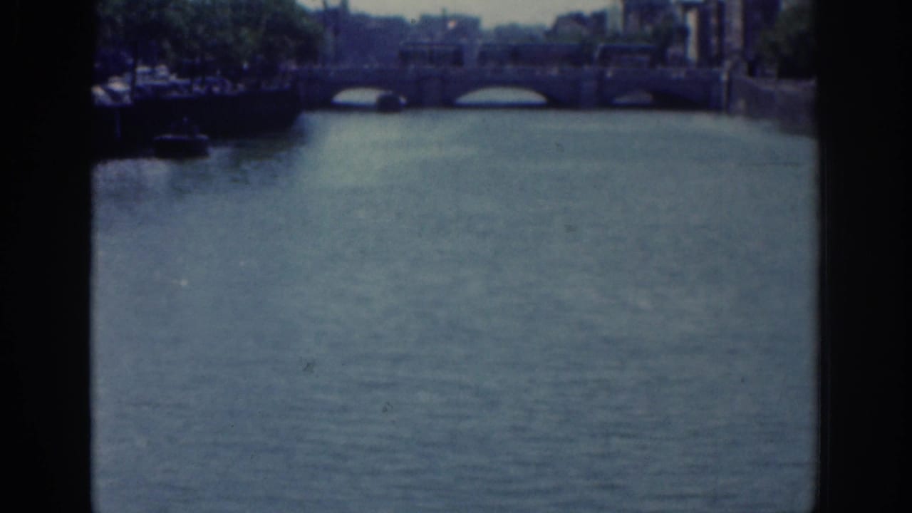 1961 Dublin home-movie frame with a closer view of river water, bridge arches, tree cover, and riverside buildings.