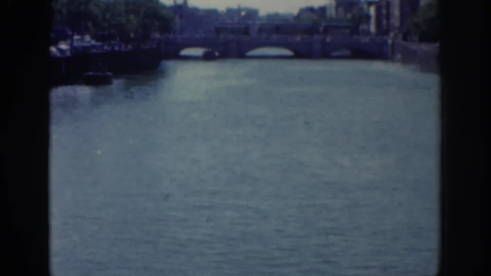 1961 Dublin home-movie frame with a closer view of river water, bridge arches, tree cover, and riverside buildings.