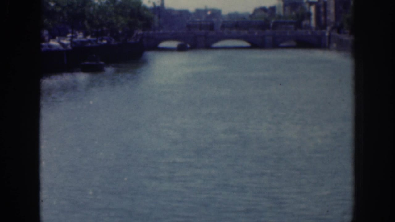 1961 Dublin 8mm frame showing a broad river surface leading toward a multi-arch stone bridge and shaded urban banks.