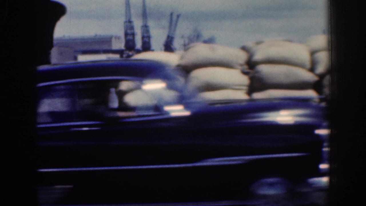 1961 Dublin home-movie frame showing a dark vehicle or cart loaded with sacks moving past a bridge railing, with port cranes in the background.