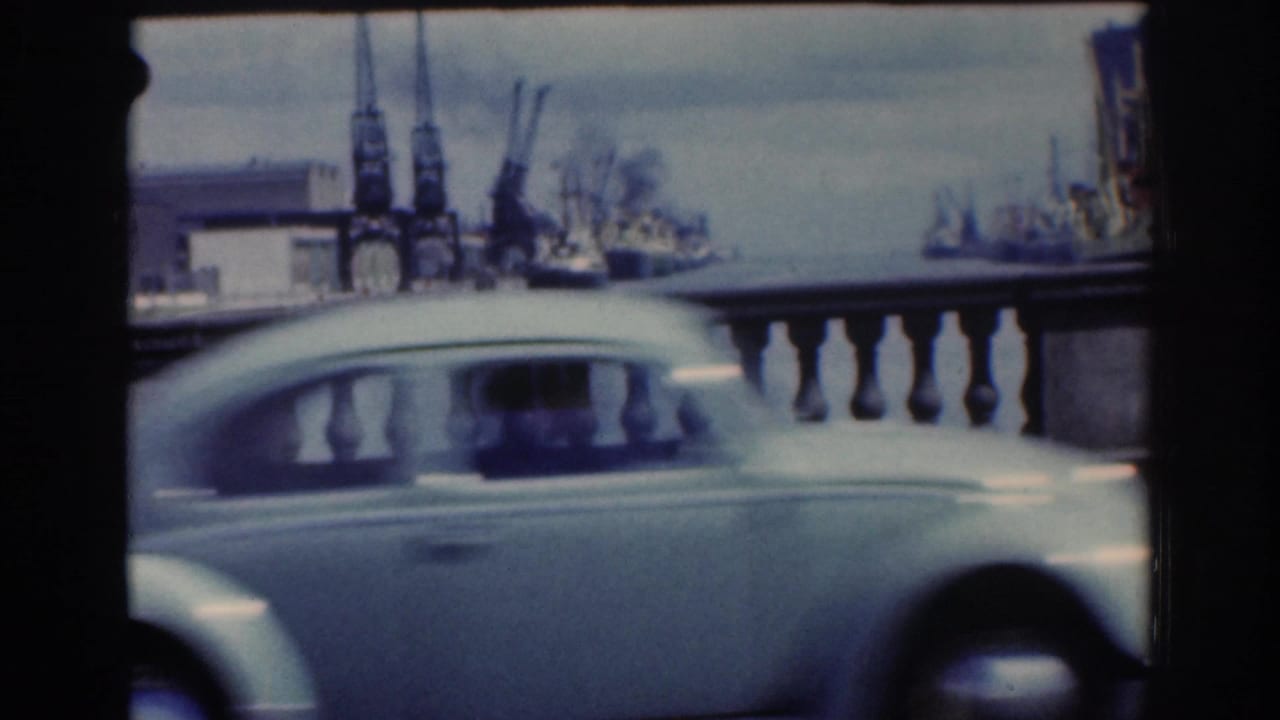 Light-colored small car passes in front of a stone railing with port cranes and ships behind.