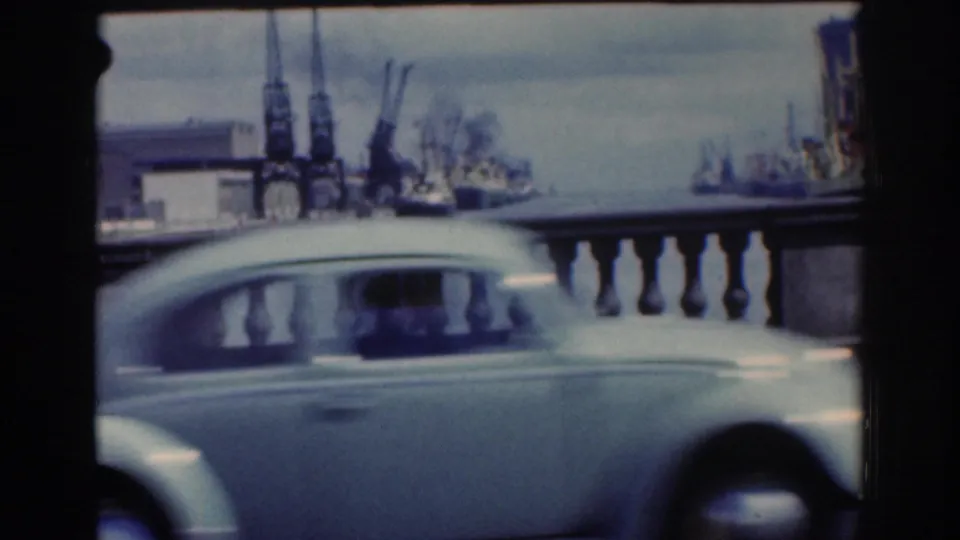Light-colored small car, likely a period compact, drives along the Dublin waterfront with cranes and ships visible beyond the railing.
