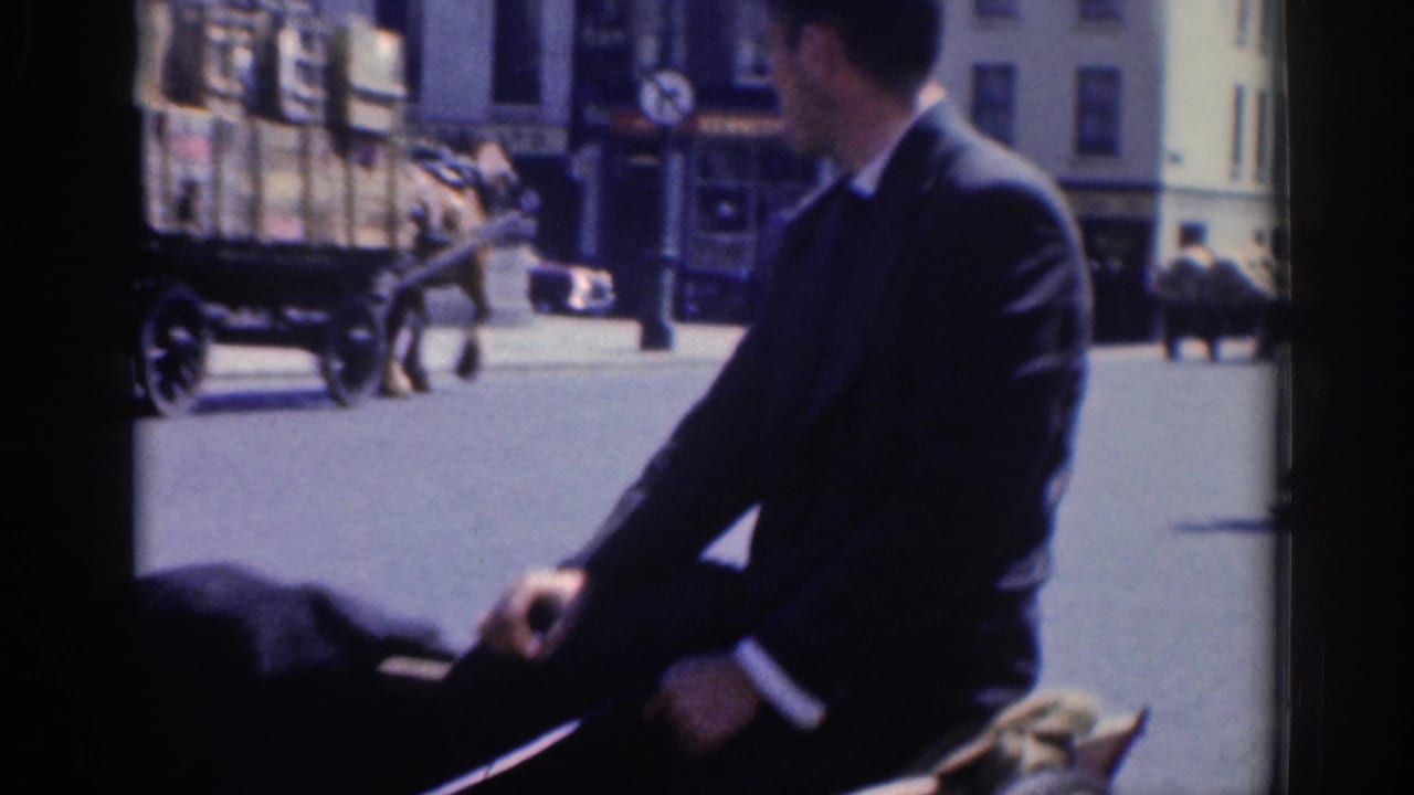 A suited man, likely with a carriage, sits beside a horse on a Dublin street with buildings and pedestrians behind him.