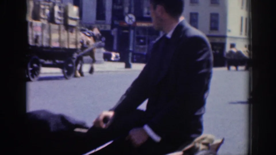 A suited man, likely with a carriage, sits beside a horse on a Dublin street with buildings and pedestrians behind him.