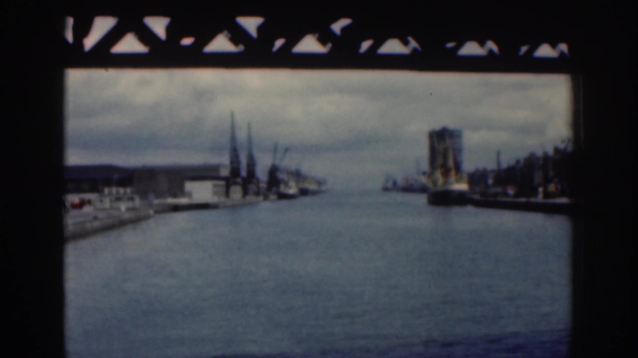 1961 Dublin river or canal view framed beneath a bridge, with industrial waterfront buildings, cranes, and a large vessel along the right bank.
