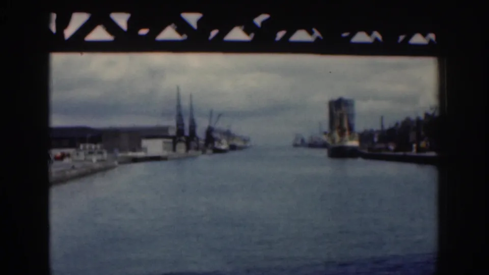 1961 Dublin river or canal view framed beneath a bridge, with industrial waterfront buildings, cranes, and a large vessel along the right bank.
