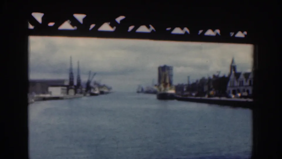 1961 Dublin riverfront framed by a bridge, with cranes and low port buildings on the left and a large ship or tall structure on the right bank.