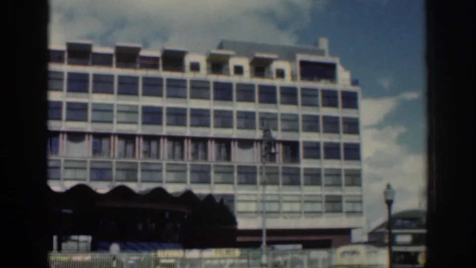 Large modernist multi-story building in Dublin, 1961, viewed from a street-level moving home-movie perspective.