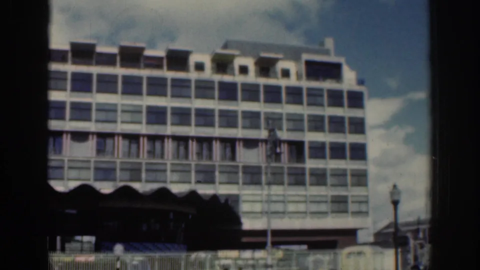1961 Dublin street view showing a large gridded building facade, dark awning line, and street lamps from 8mm footage.