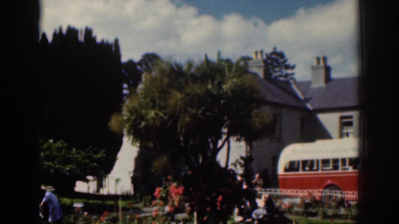 1961 Dublin home-movie frame showing a red and white bus near a garden, with trees, flowers, pedestrians, and houses behind.