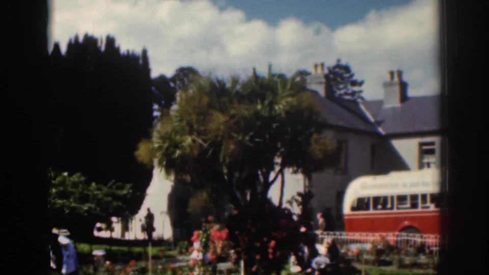 1961 Dublin frame with a red and white bus at a garden-side stop, pedestrians in period dress, trees, flowers, and houses in back.