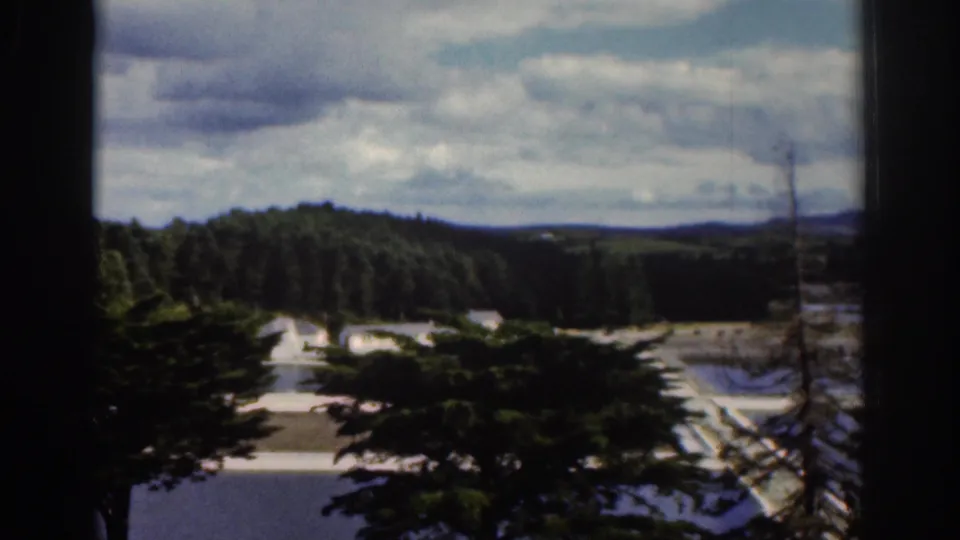 1961 Dublin, Ireland home-movie frame looking through a window toward wooded hills, pale roads or paths, and a clouded sky.