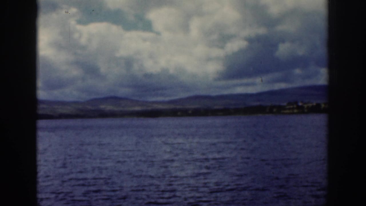 1961 Dublin, Ireland frame showing open water, a low distant shore, and mountains under a dramatic cloudy sky.