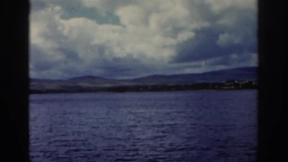 1961 Dublin, Ireland frame showing open water, a low distant shore, and mountains under a dramatic cloudy sky.