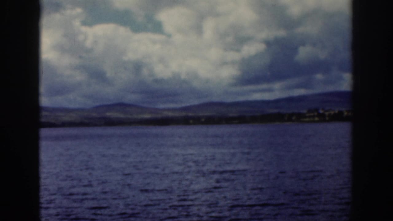 1961 Dublin travel footage showing water, distant hills, shoreline buildings, and a dark tree trunk before the ruin view.