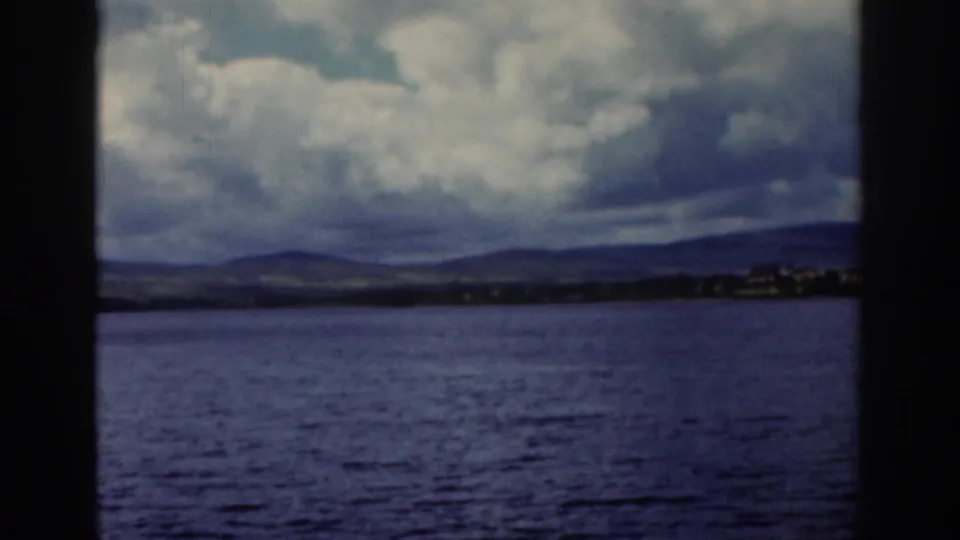 1961 Dublin travel footage showing water, distant hills, shoreline buildings, and a dark tree trunk before the ruin view.