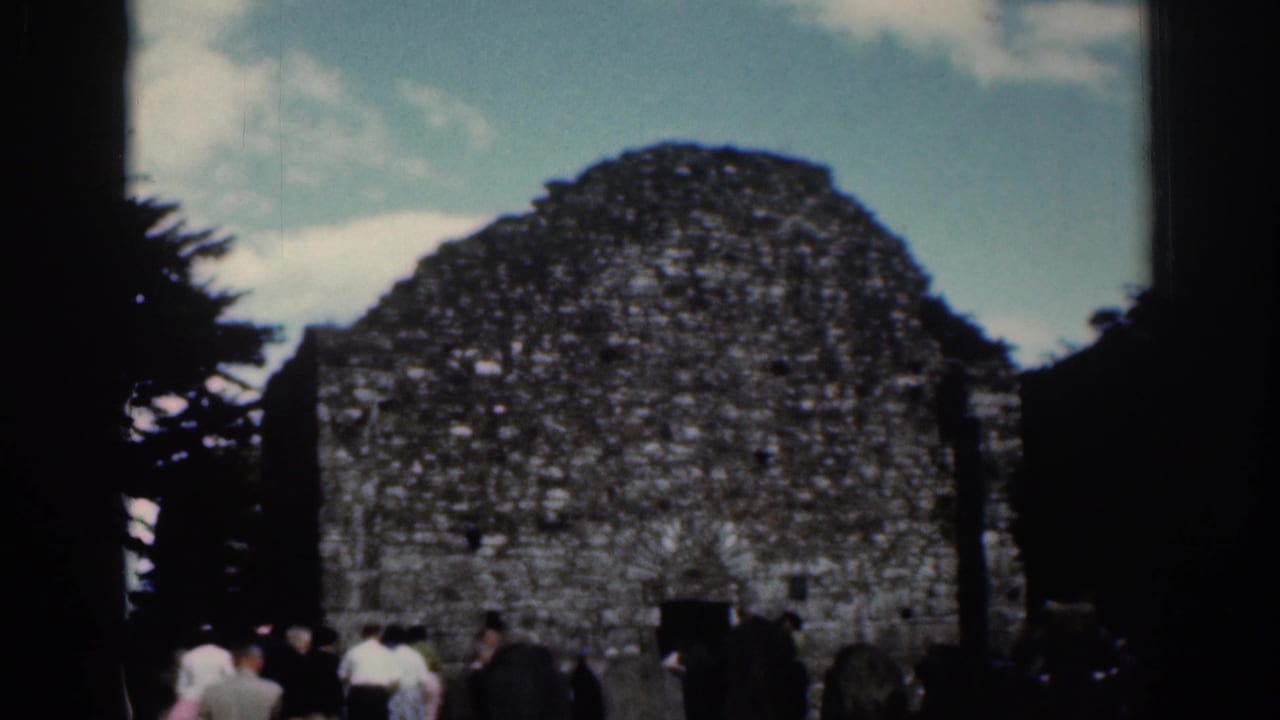 Tourists stand near a weathered stone ruin framed by dark trees and blue sky.