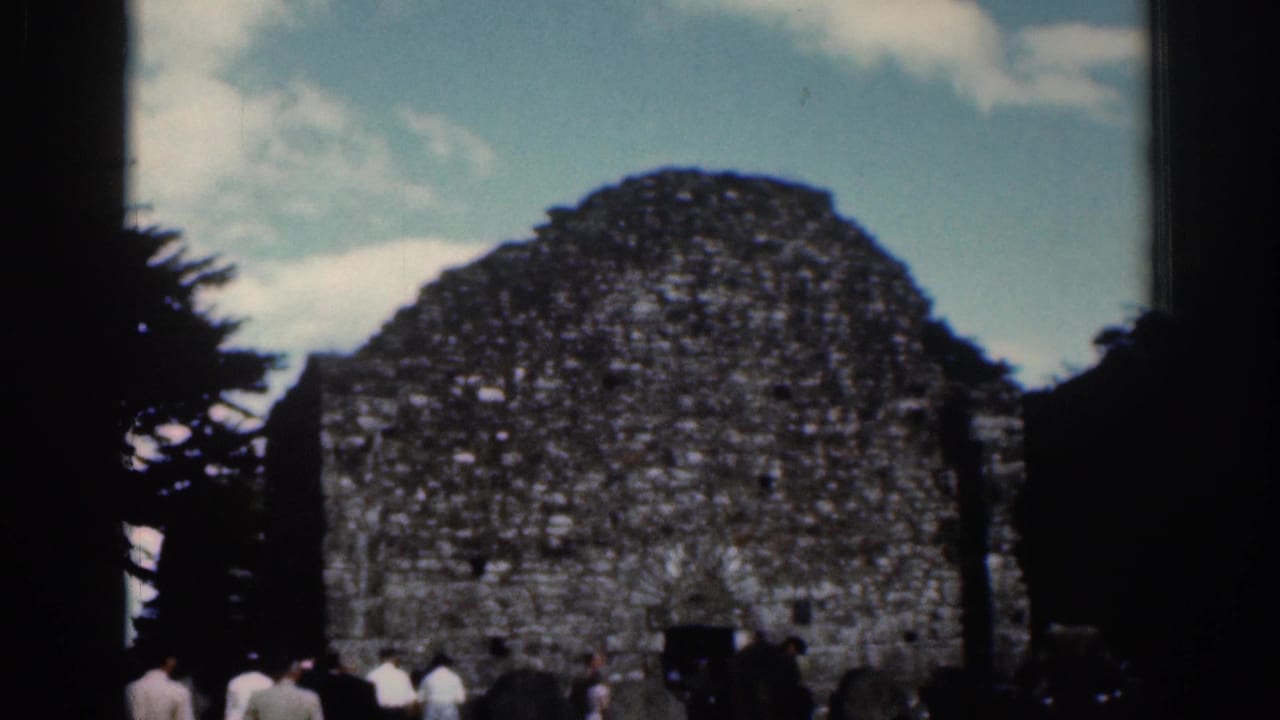 1961 Dublin frame centered on a weathered stone structure with arched openings, tourists, trees, and pale blue sky.