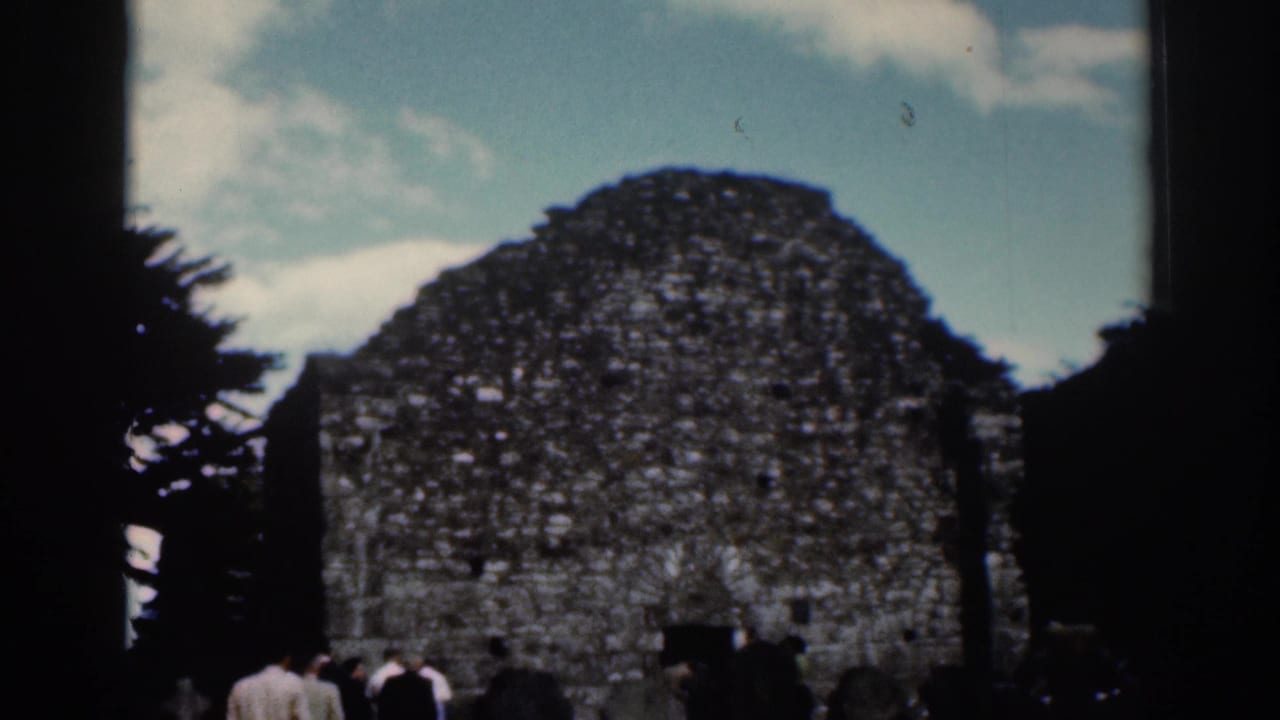 1961 Dublin home-movie view of the stone ruin, small visitor group, dark tree trunks, and scattered clouds.