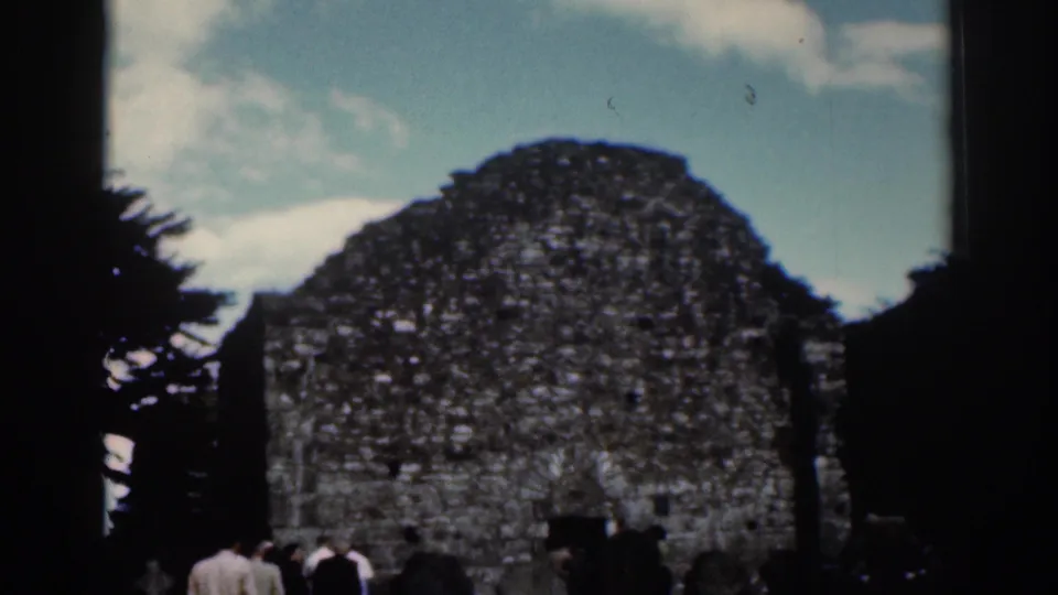 1961 Dublin home-movie view of the stone ruin, small visitor group, dark tree trunks, and scattered clouds.
