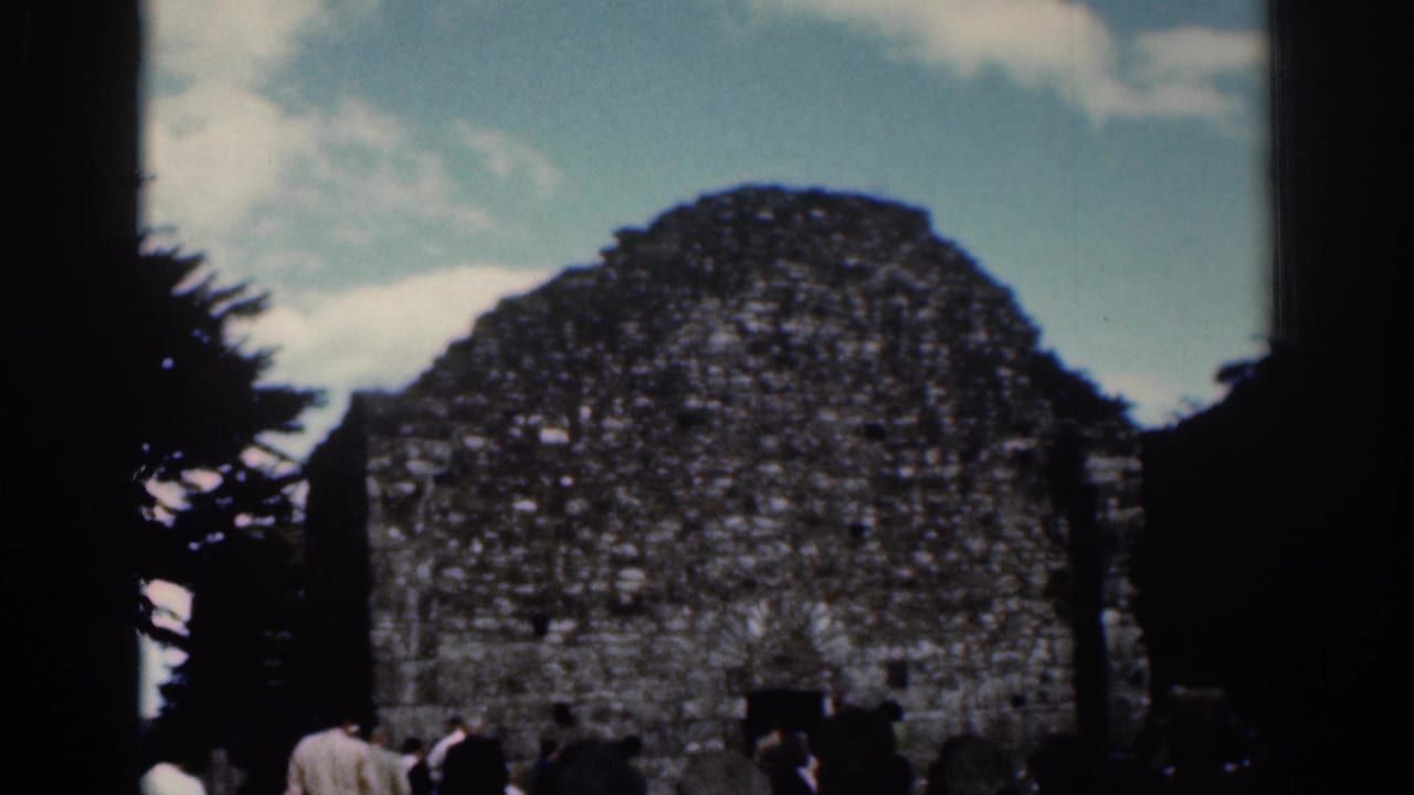 1961 Dublin 8mm frame showing visitors beside weathered stone ruins, a dark doorway, trees, and cloudy sky.