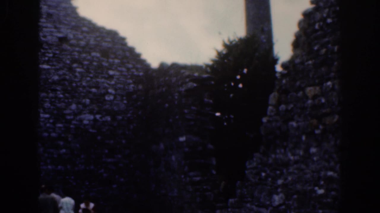 1961 Dublin 8mm frame with shadowed stone walls, a tree trunk, foliage, and distant visitors at the base of the ruin.