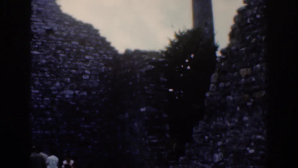 1961 Dublin 8mm frame with shadowed stone walls, a tree trunk, foliage, and distant visitors at the base of the ruin.