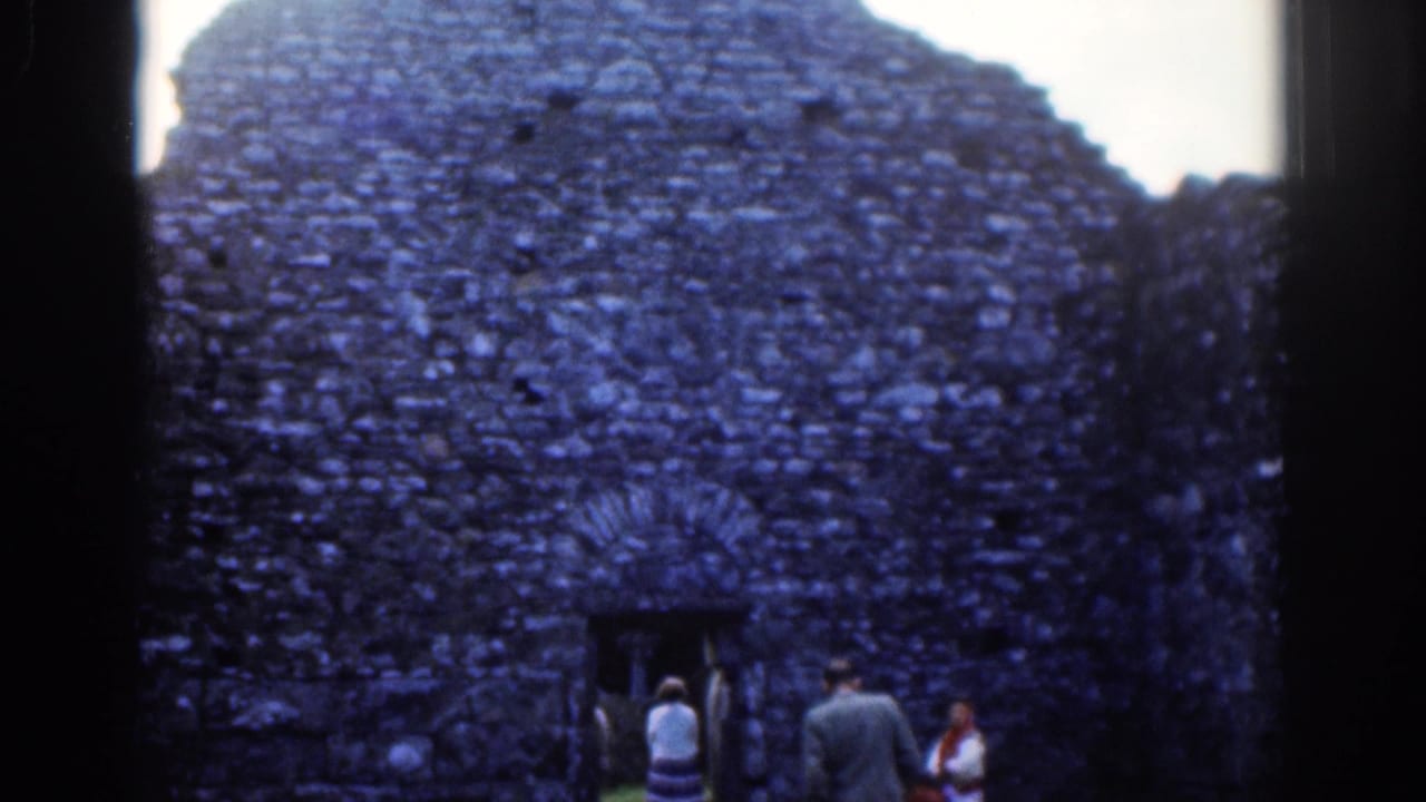 Adults stand near a rough stone wall with an arched doorway at a ruin.