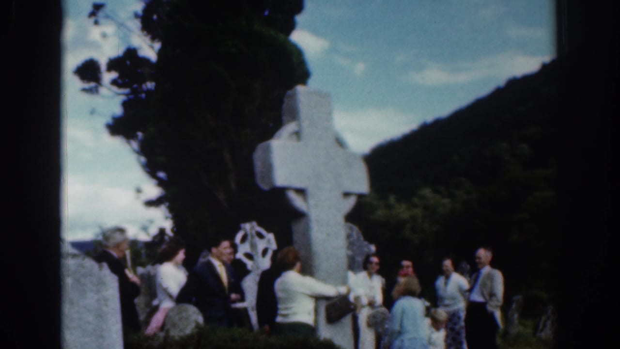Adults in 1961 Dublin stand near a large Celtic cross with trees and hills behind them.