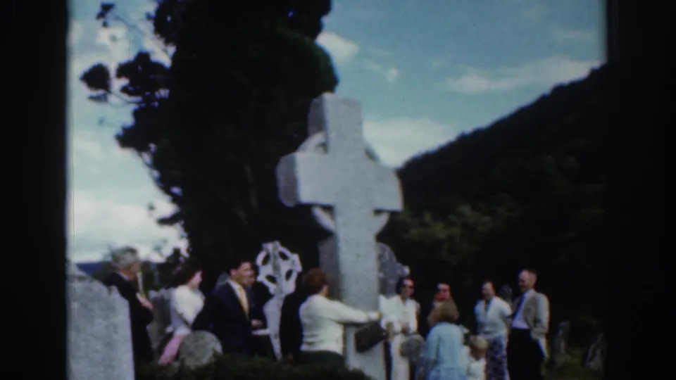 Adults in 1961 Dublin stand near a large Celtic cross with trees and hills behind them.