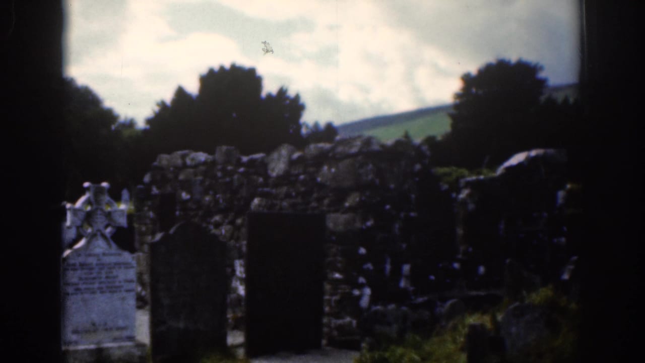 Gravestones and a Celtic cross in a churchyard with a stone wall and hills behind.
