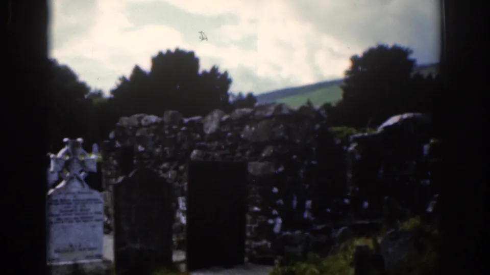 1961 Dublin-area home-movie view of a churchyard with weathered graves, a pale Celtic cross, rough stonework, trees, and green hills.