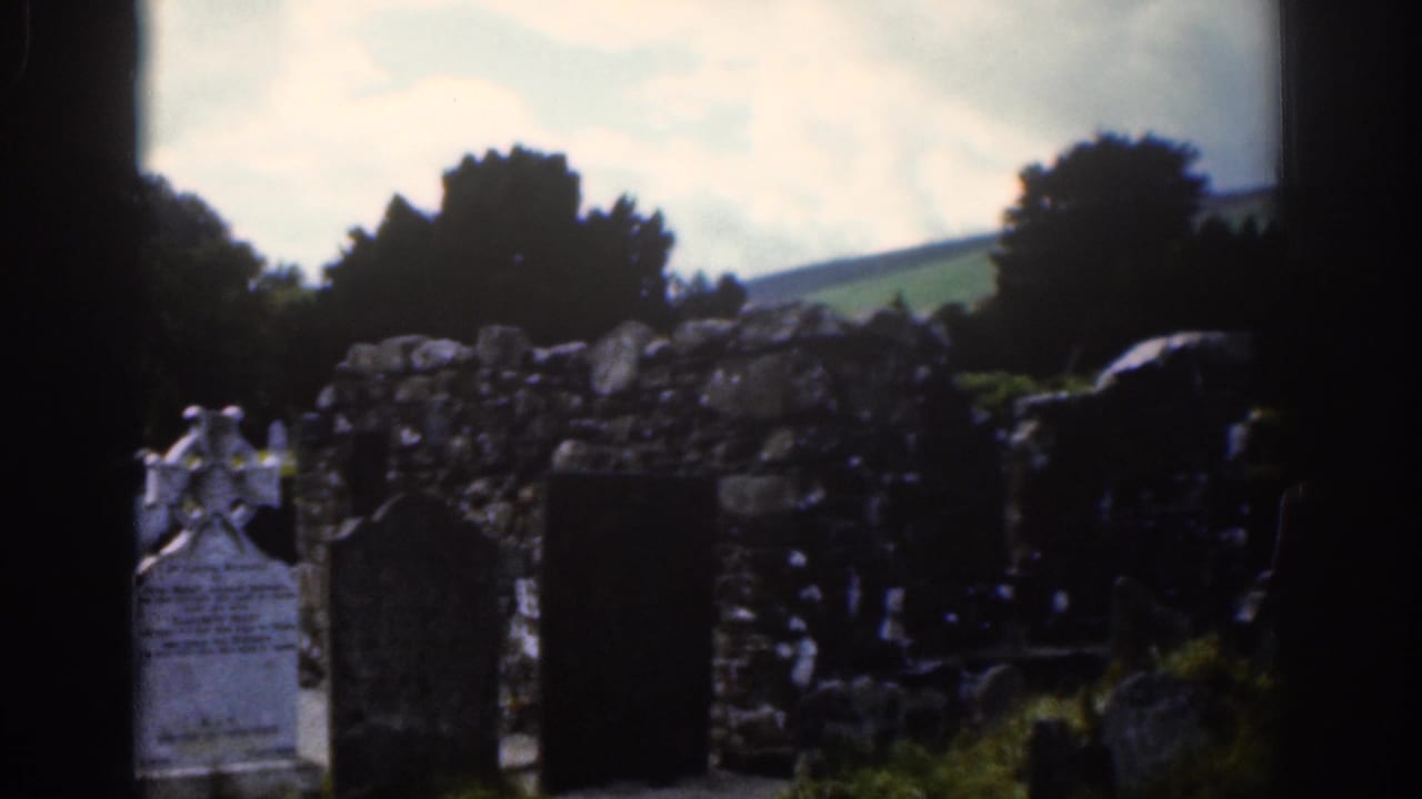 Churchyard graves and a Celtic cross beside rough stonework under a cloudy sky.