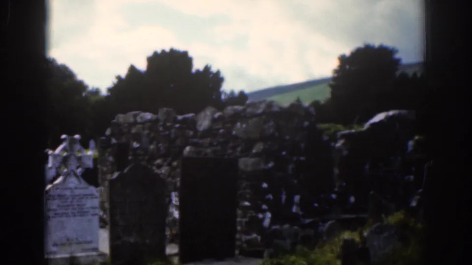 1961 Dublin-area 8mm frame showing cemetery markers, a Celtic cross, rough stone masonry, dark trees, and hills beneath cloud cover.