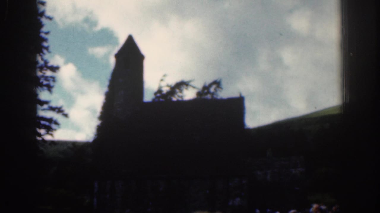 1961 Dublin-area home-movie frame with a church tower and roofline silhouetted against bright clouds, with trees and hillside nearby.